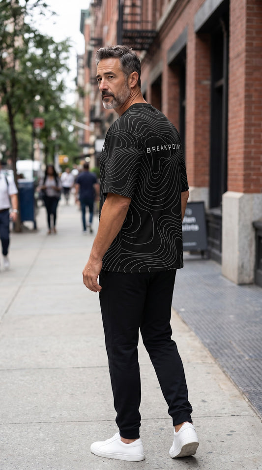 Man wearing a black BREAKPOINT Sportswear Courtside Tee with white topographic line graphics on a city sidewalk.